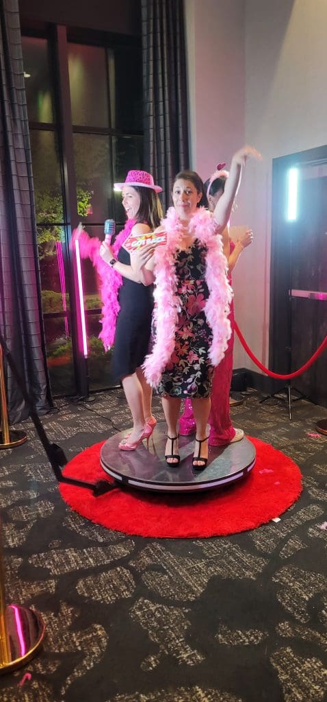 Three women dressed up with pink feather boas and props, posing on a photo booth platform at an event.
