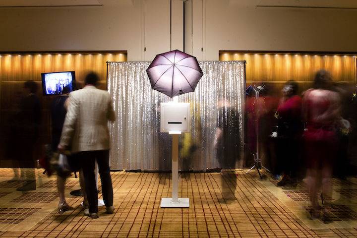 Photo booth setup with a pink umbrella light on a stand in front of a silver sequin backdrop; guests mingle in the blurred background at an event.