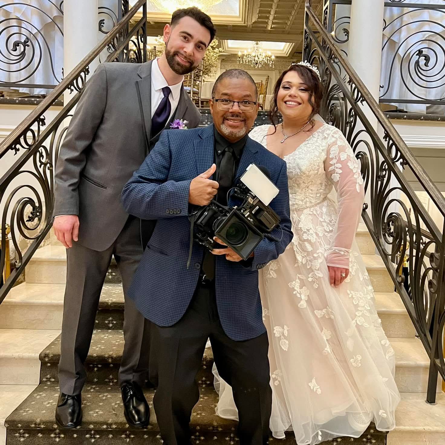 Photographer in blue blazer gives thumbs up with wedding couple on a grand staircase in formal attire (groom in gray suit, bride in lace gown) and chandelier background.