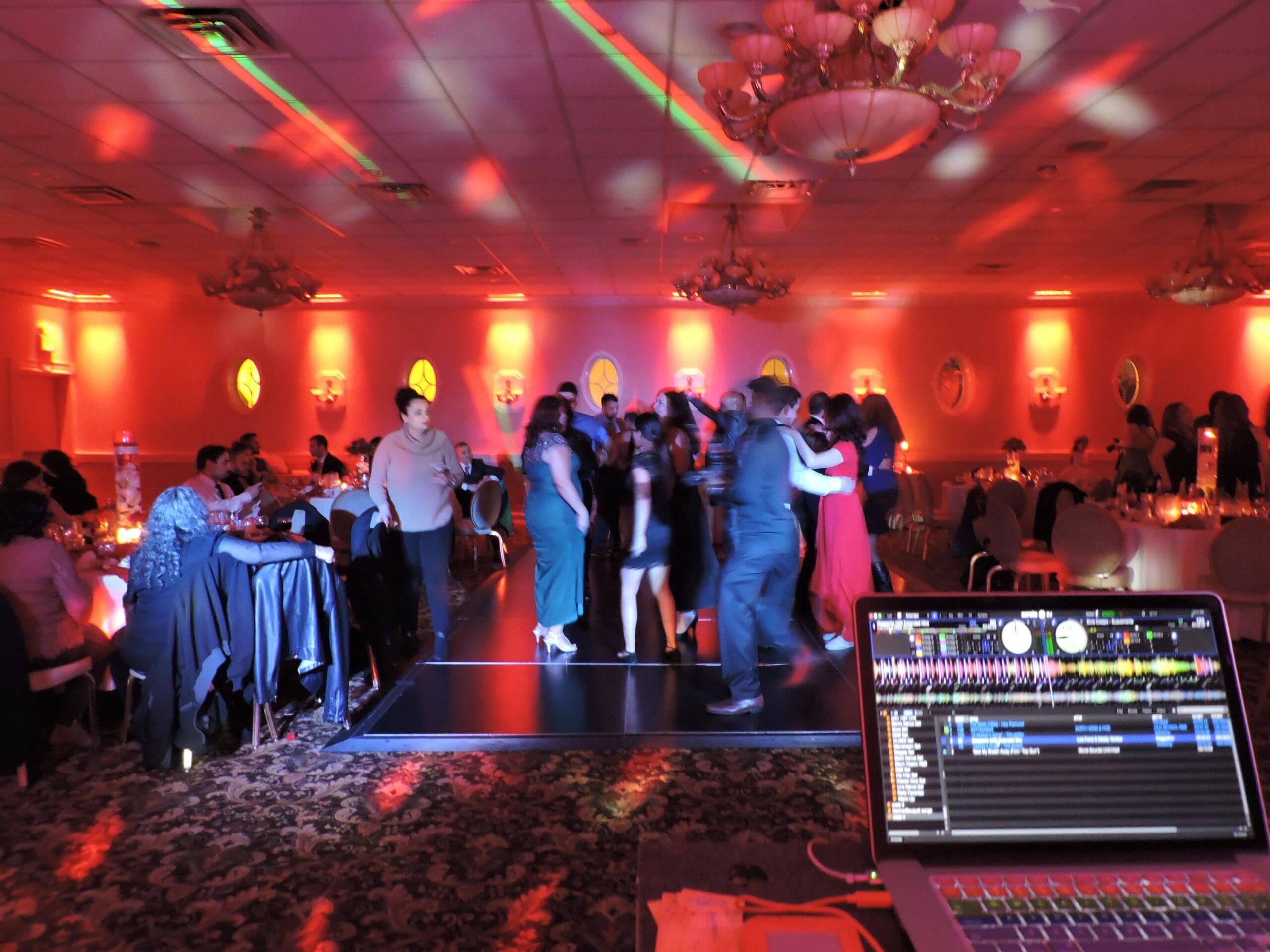 A crowded ballroom with guests dancing under red and purple uplighting and chandeliers, DJ equipment visible in the foreground.